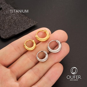 Puede incluir: Cuatro pendientes de aro de titanio en tonos dorados y plateados. Los pendientes son de varios tama&ntilde;os y se muestran en una mano. La palabra "TITANIUM" est&aacute; en la parte superior izquierda de la imagen. La marca "OUFER BODY JEWELRY" est&aacute; en la parte inferior derecha.