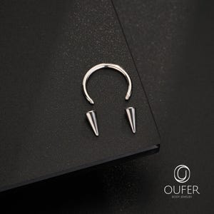 May include: A silver-colored circular barbell body jewelry piece featuring a curved metal bar with threaded ends and two detachable conical spike attachments. The metal components have a polished finish and are displayed with the OUFER BODY JEWELRY logo.