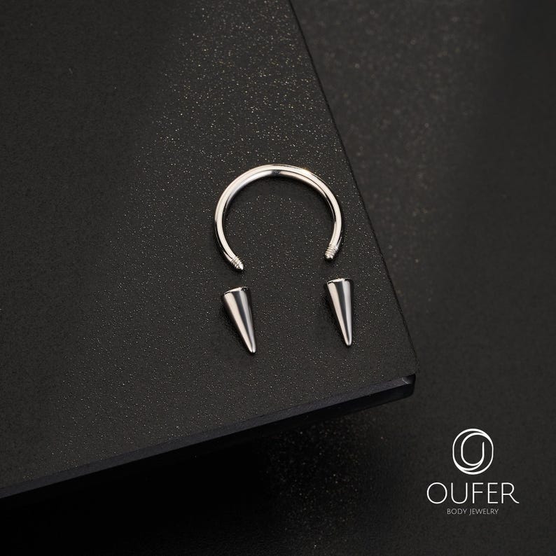 May include: A silver-colored circular barbell body jewelry piece featuring a curved metal bar with threaded ends and two detachable conical spike attachments. The metal components have a polished finish and are displayed with the OUFER BODY JEWELRY logo.