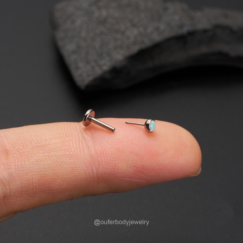 Op de afbeelding: Een paar lichaamssieraden van chirurgisch staal met een kleine opaal-edelsteen. De studs worden op een vinger getoond.