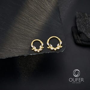 Op de afbeelding: Twee goudkleurige lichaamsjuwelen ringen, elk met een cirkelvormig ontwerp. De ringen zijn versierd met kleine, witte, bladvormige versieringen. De sieraden worden gepresenteerd op een donkere, getextureerde ondergrond.