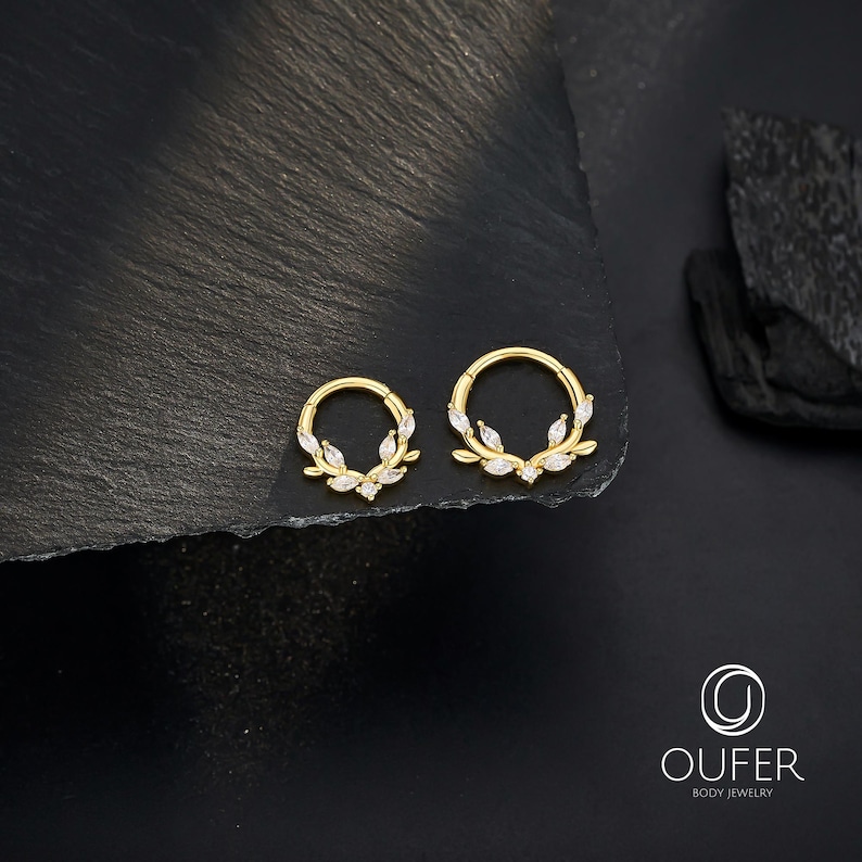 Op de afbeelding: Twee goudkleurige lichaamsjuwelen ringen, elk met een cirkelvormig ontwerp. De ringen zijn versierd met kleine, witte, bladvormige versieringen. De sieraden worden gepresenteerd op een donkere, getextureerde ondergrond.