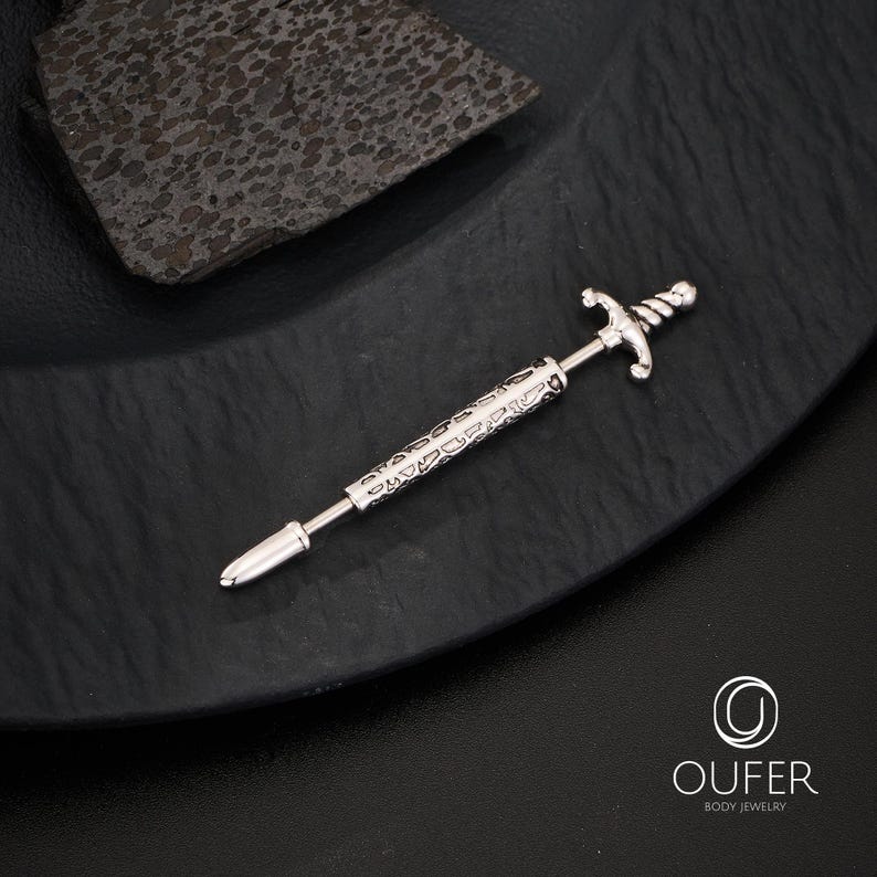 K&ouml;nnte beinhalten: Ein silberfarbener Piercing-Barbell mit Schwertdesign. Der Barbell hat einen detaillierten Schwertknauf und eine Klinge mit ge&auml;tzten Mustern entlang des Schafts. Der Schmuck wird auf einer schwarzen, strukturierten Oberfl&auml;che pr&auml;sentiert.