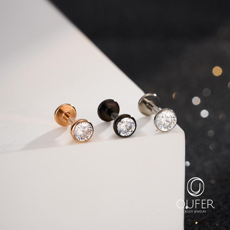 Puede incluir: Se muestran tres piezas de joyer&iacute;a corporal. Una es de oro rosa, otra negra y otra plateada. Cada pieza presenta una peque&ntilde;a gema redonda y transparente. La joyer&iacute;a se encuentra sobre un fondo blanco y negro.