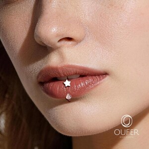 May include: Close-up of a person's lips with two pieces of body jewelry. A small, white star-shaped stud is on the upper lip, and a teardrop-shaped stud is on the lower lip. The lips are a natural, reddish-brown color.