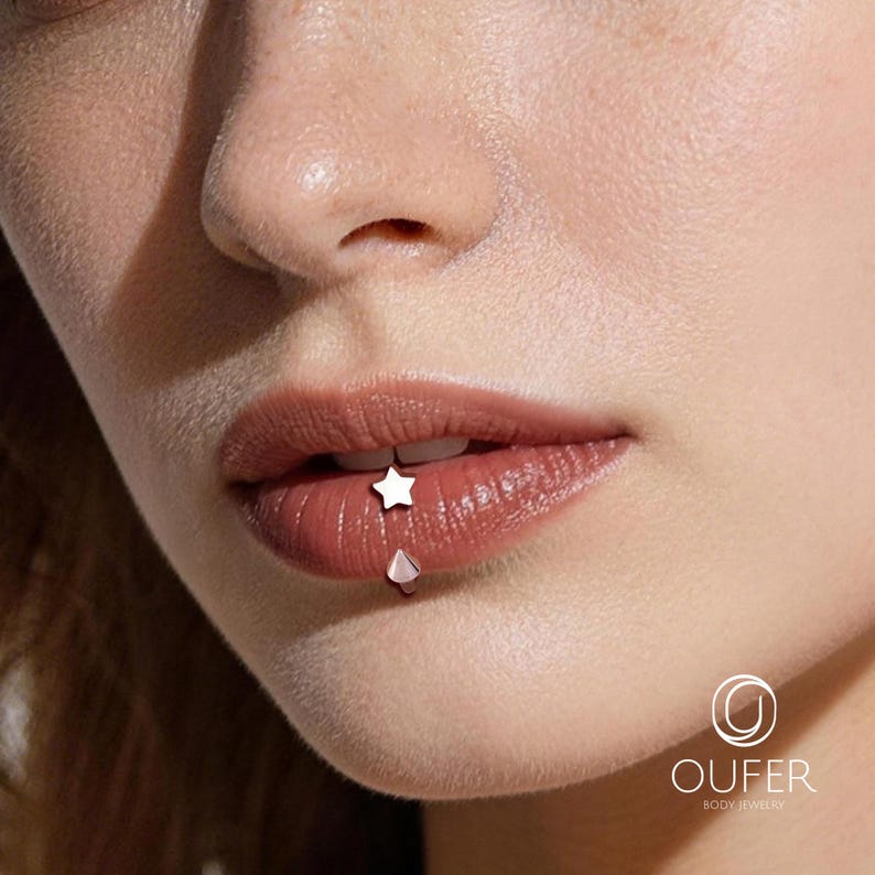 May include: Close-up of a person's lips with two pieces of body jewelry. A small, white star-shaped stud is on the upper lip, and a teardrop-shaped stud is on the lower lip. The lips are a natural, reddish-brown color.