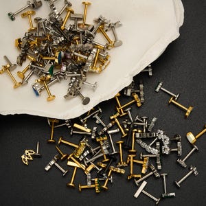 Puede incluir: Una colecci&oacute;n de pendientes y piercings para labios surtidos en tonos dorados y plateados, algunos con detalles de piedras preciosas. Los pendientes est&aacute;n esparcidos sobre una superficie blanca texturizada y un fondo negro.