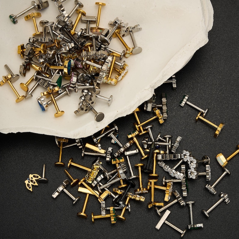 Puede incluir: Una colecci&oacute;n de pendientes y piercings para labios surtidos en tonos dorados y plateados, algunos con detalles de piedras preciosas. Los pendientes est&aacute;n esparcidos sobre una superficie blanca texturizada y un fondo negro.