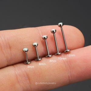 Op de afbeelding: Een set van zes zilveren barbell oorbellen met opaalstenen. De oorbellen zijn gerangschikt op grootte, van 5 mm tot 12 mm. De oorbellen bevinden zich op een vinger, met de afmetingen eronder aangegeven.