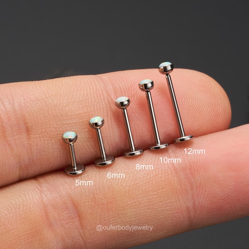 Op de afbeelding: Een set van zes zilveren barbell oorbellen met opaalstenen. De oorbellen zijn gerangschikt op grootte, van 5 mm tot 12 mm. De oorbellen bevinden zich op een vinger, met de afmetingen eronder aangegeven.