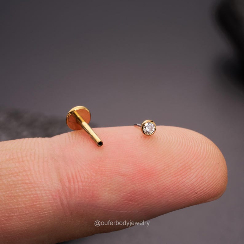 May include: Two gold body jewelry pieces, one is a straight barbell with a flat end and the other is a small stud with a clear gemstone.