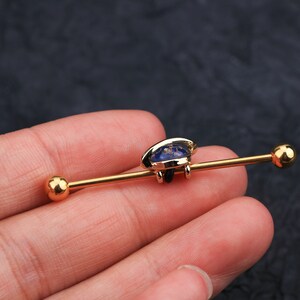 Puede incluir: Pendiente industrial barbell de color dorado con un dise&ntilde;o azul y dorado. El pendiente presenta una barra recta con una bola en cada extremo. El centro tiene un elemento decorativo con un dise&ntilde;o azul y dorado.