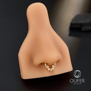 Op de afbeelding: Een goudkleurige septumring met een bladeren design, versierd met heldere edelstenen. De sieraden worden getoond op een neusmodel, om de pasvorm en stijl van het product te laten zien. De ring is lichaamsjuwelen.