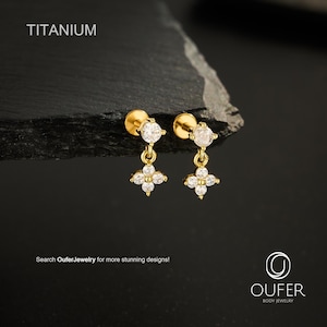 Puede incluir: Dos pendientes de botón de titanio en tono dorado con un colgante de flor de cuatro pétalos engastado con piedras redondas transparentes. Cada pendiente tiene una piedra clara en el poste y un colgante floral. Texto TITANIUM y OUFER.