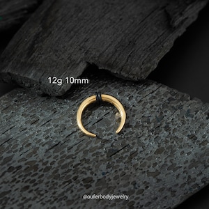May include: Gold 10mm 12 gauge septum ring with black o-rings. The ring is on a black background.