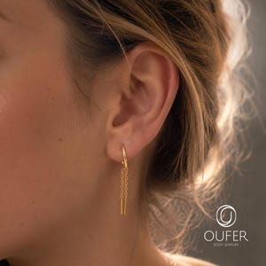 May include: A gold hoop earring featuring two delicate dangling chains that end in small, straight metal bars. The earring is worn in the earlobe, showcasing a polished finish and a minimalist design that hangs vertically from the circular hoop.