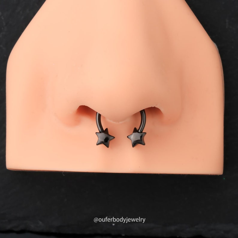 May include: A close-up of a black nose ring with star-shaped ends. The ring is worn in a pierced nose.