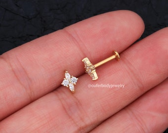 May include: Gold body jewelry with a small flower design featuring clear cubic zirconia stones. The jewelry is shown on a person's hand.