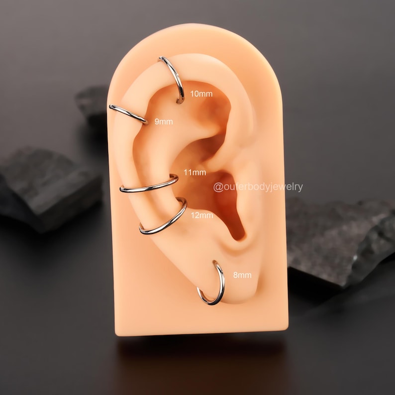 Puede incluir: Primer plano de una oreja modelo con cinco aros de plata de diferentes tama&ntilde;os. Los aros est&aacute;n etiquetados con su di&aacute;metro en mil&iacute;metros: 8 mm, 10 mm, 9 mm, 11 mm y 12 mm. El texto "@outerbodyjewelry" tambi&eacute;n es visible en la imagen.