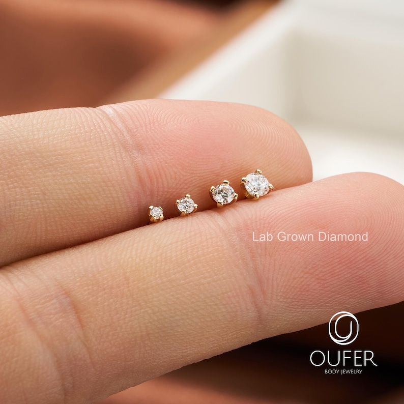 Puede incluir: Cuatro pendientes de bot&oacute;n dorados con diamantes redondos y transparentes cultivados en laboratorio de diferentes tama&ntilde;os se muestran en un dedo. Los pendientes tienen un dise&ntilde;o sencillo y cl&aacute;sico. El texto "Lab Grown Diamond" es visible.