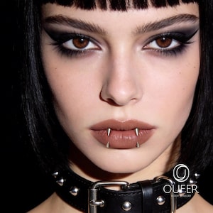 May include: A silver-toned metal lip piercing featuring two sharp, conical spikes positioned vertically on the lower lip. The jewelry is worn by an individual with dark hair and a black studded leather choker. The OUFER BODY JEWELRY logo is visible.