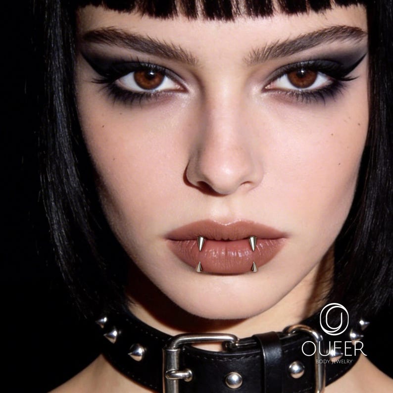 May include: A silver-toned metal lip piercing featuring two sharp, conical spikes positioned vertically on the lower lip. The jewelry is worn by an individual with dark hair and a black studded leather choker. The OUFER BODY JEWELRY logo is visible.
