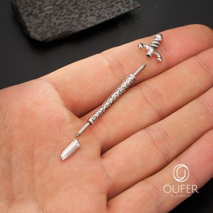 K&ouml;nnte beinhalten: Ein zerlegtes silberfarbenes K&ouml;rperschmuckst&uuml;ck mit einem schwertartigen Design. Das Schmuckst&uuml;ck besteht aus mehreren Teilen, darunter ein dekorativer Schaft mit einem ge&auml;tzten Muster und eine schwertf&ouml;rmige Spitze. Der Schmuck wird vor einem neutralen Hintergrund pr&auml;sentiert.
