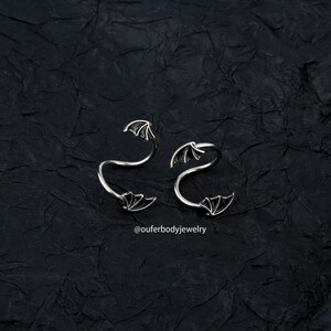 Peut inclure: Deux anneaux en spirale argent&eacute;s avec des motifs d'ailes de chauve-souris. Les anneaux sont sur un fond textur&eacute; noir. Le texte "@ouferbodyjewelry" est en bas de l'image.