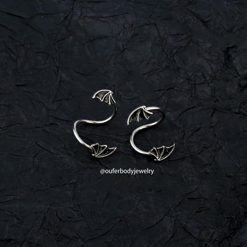 Peut inclure: Deux anneaux en spirale argent&eacute;s avec des motifs d'ailes de chauve-souris. Les anneaux sont sur un fond textur&eacute; noir. Le texte "@ouferbodyjewelry" est en bas de l'image.