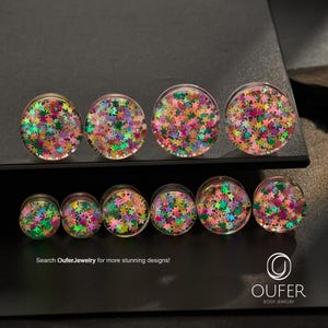 Puede incluir: Diez expansores de oreja circulares de varios tamaños dispuestos en dos filas. Cada expansor es de material transparente relleno de pequeñas purpurinas en forma de estrella multicolor en tonos rosa, verde, azul y dorado. Texto: Search OuferJewelry.