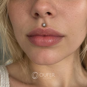 Puede incluir: Un piercing de filtrum en tono plateado con un diseño circular que presenta una gema transparente central rodeada de pequeñas cuentas metálicas. La joya se sitúa sobre el labio superior. El texto OUFER BODY JEWELRY es visible abajo.
