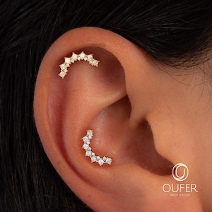 May include: Close-up of an ear adorned with two pieces of jewelry. The upper piece is a curved, gold-toned earring with multiple clear, square-cut stones. The lower piece is a similar earring with a curved design and clear stones. The jewelry is from Oufer Body Jewelry.