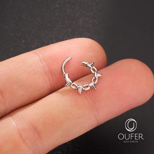 Op de afbeelding: Een zilverkleurige scharnierende oorring met een kettingachtig ontwerp en kleine, puntige spikes. De oorring is open en toont het scharniermechanisme. De achtergrond is een vage donkere kleur.