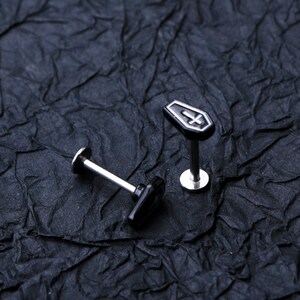 May include: Two black and silver body jewelry pieces. One piece is a coffin-shaped stud with a silver cross. The other piece is a black stud with a silver post.
