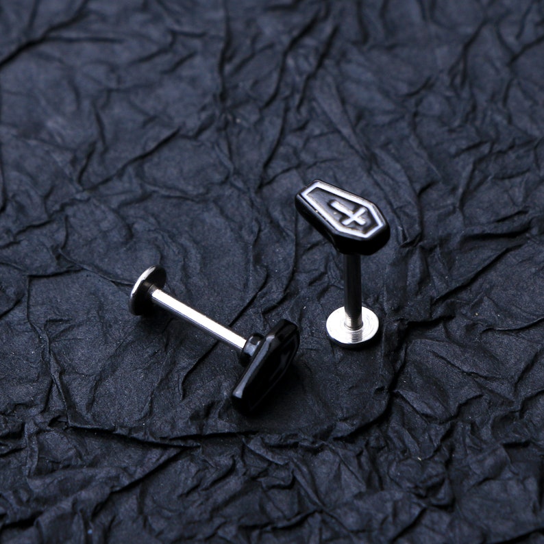 May include: Two black and silver body jewelry pieces. One piece is a coffin-shaped stud with a silver cross. The other piece is a black stud with a silver post.