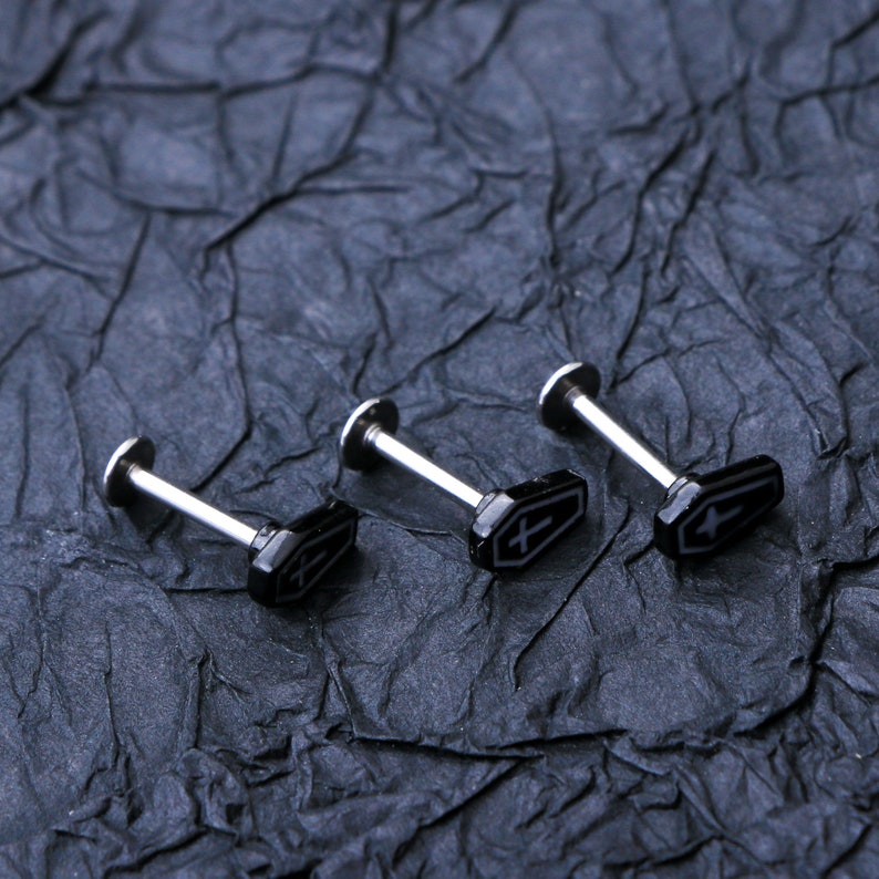 May include: Three silver body jewelry pieces with black coffin-shaped charms. Each charm has a small white cross on it.