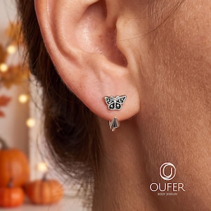 May include: A silver-toned earring featuring a butterfly-shaped stud with intricate black detailing on the wings, paired with a small, pointed spike charm that hangs from the back of the earlobe, creating a layered and dimensional appearance.