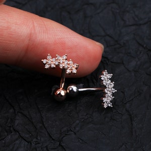May include: Two silver and rose gold body jewellery pieces with a flower design. Each piece has a small ball on one end and a cluster of small flowers on the other end. The flowers are set with clear rhinestones.
