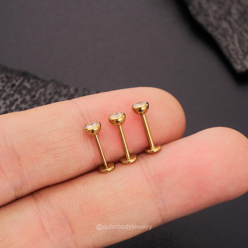 May include: Three gold-toned lip rings with clear crystal accents. The rings are arranged in a row, with the largest ring on the left and the smallest ring on the right.