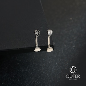 Puede incluir: Dos piezas de joyería corporal tipo barra curva con engastes en forma de lágrima. Una pieza tiene una piedra negra y la otra una piedra blanca transparente. Ambas tienen un acabado metálico plateado y una base circular plana. Se lee OUFER.