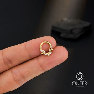 Op de afbeelding: Een goudkleurige klapcreool-oorbel met een bladerenontwerp. De oorbel heeft kleine, heldere, marquise-geslepen stenen gerangschikt in een kranspatroon. De sieraden worden gepresenteerd tegen een donkere achtergrond.