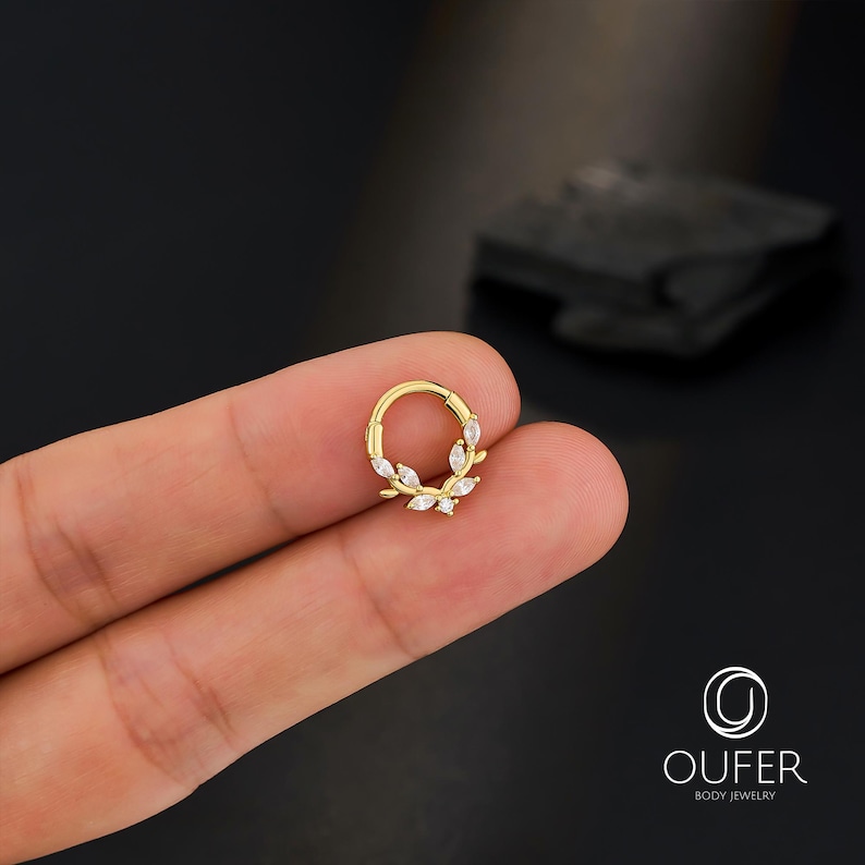 Op de afbeelding: Een goudkleurige klapcreool-oorbel met een bladerenontwerp. De oorbel heeft kleine, heldere, marquise-geslepen stenen gerangschikt in een kranspatroon. De sieraden worden gepresenteerd tegen een donkere achtergrond.