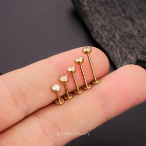 May include: A set of six gold-toned body jewelry pieces with clear gem accents. The jewelry is arranged in a line, with the largest piece at the top and the smallest piece at the bottom.