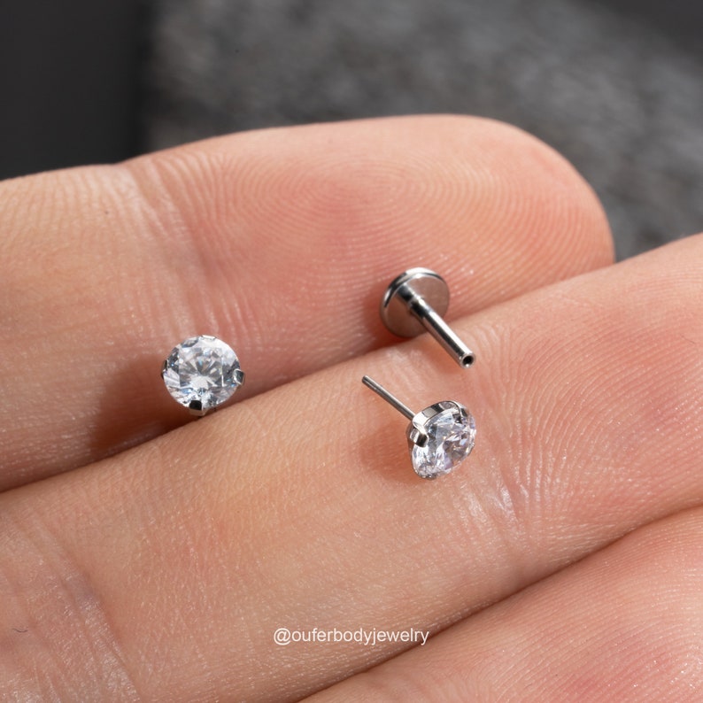 Puede incluir: Dos piezas de joyer&iacute;a corporal plateadas con gemas redondas transparentes. La joyer&iacute;a est&aacute; en un dedo.