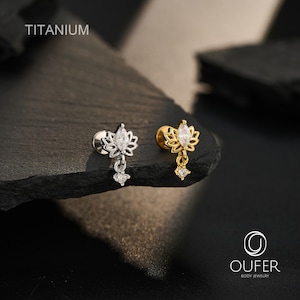 Op de afbeelding: Twee titanium lichaamsieraden met een lotusbloemontwerp, een heldere steen in marquise-slijpvorm in het midden en een kleine bungelende bedel met een ronde heldere steen. De ene knop is zilverkleurig en de andere goudkleurig. Tekst: TITANIUM.