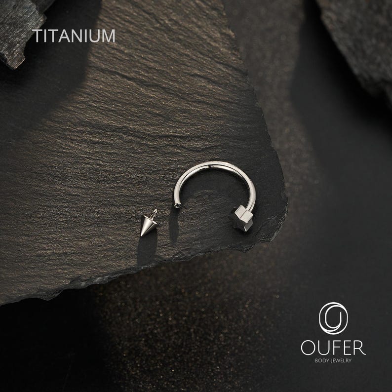 May include: A silver-colored body jewelry piece with a curved barbell design and a star-shaped end. A separate, pointed stud is also present. The word "TITANIUM" is displayed in white text. The jewelry is set against a dark, textured background.