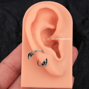 Peut inclure: Une boucle d'oreille en argent avec un motif d'aile de chauve-souris noire. La boucle d'oreille est port&eacute;e dans le cartilage sup&eacute;rieur de l'oreille.