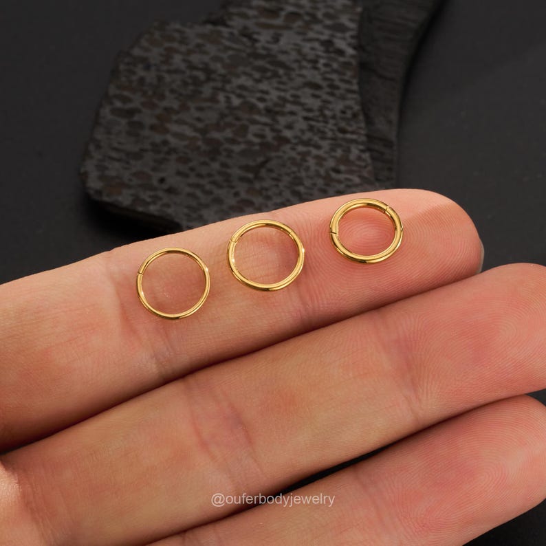 May include: Three gold seamless rings for body piercing. The rings are different sizes and are shown on a hand.