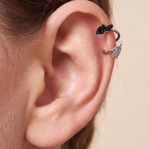 May include: A silver-toned ear cuff featuring two distinct charms attached to the band. The upper charm is a small, solid black bat silhouette, while the lower charm is a detailed silver-toned wing with textured feather-like engravings.