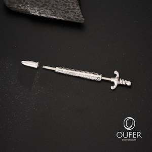 K&ouml;nnte beinhalten: Silberfarbener K&ouml;rperschmuck in Form eines Schwertes, mit strukturiertem Griff und dekorativem Knauf. Der Schmuck ist zerlegt, mit abgetrennter Spitze. Der Hintergrund ist schwarz.